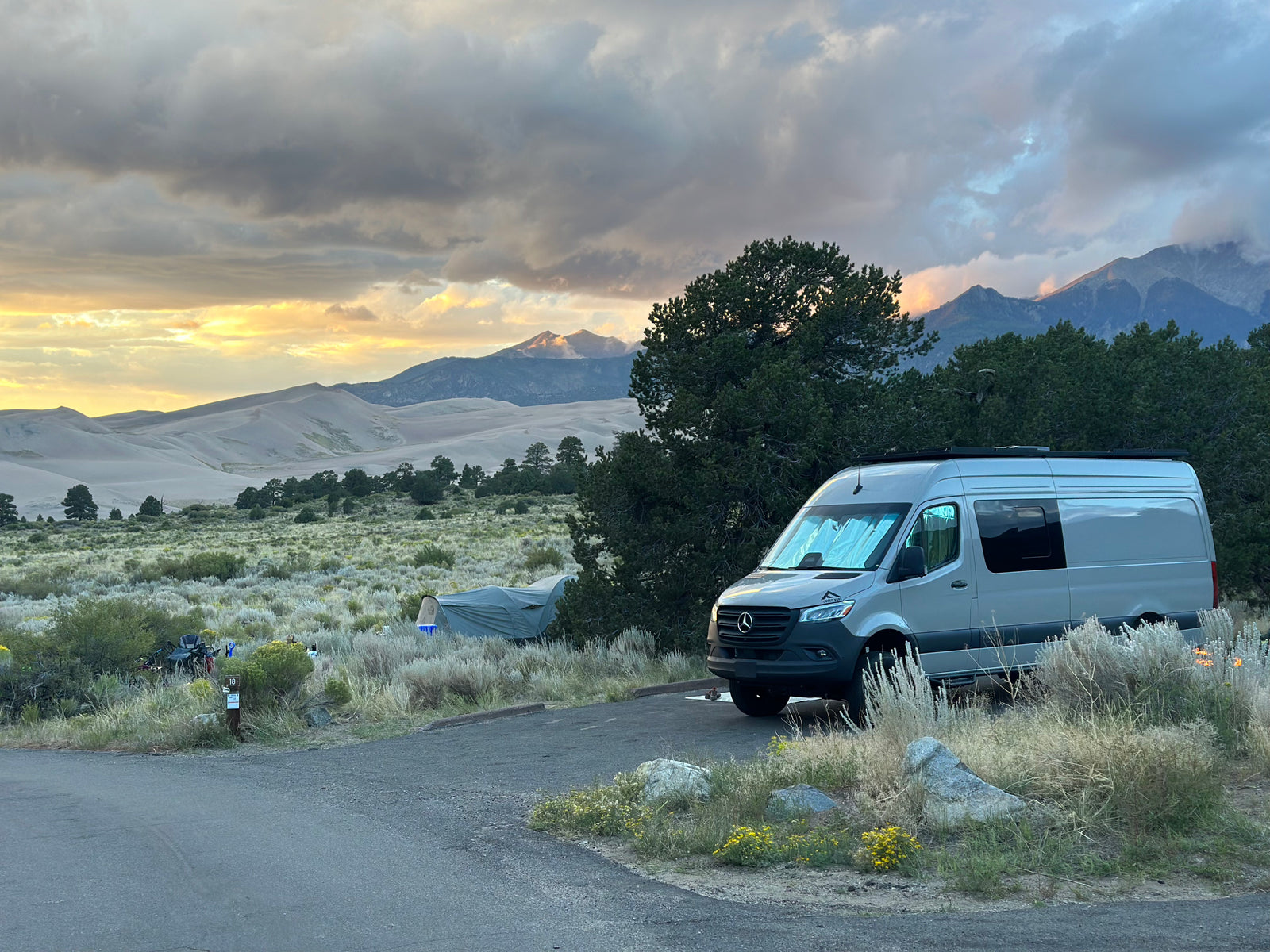RV rental camping scene: A gray camper van and tent, with majestic sand dunes and mountains under a sunset sky.