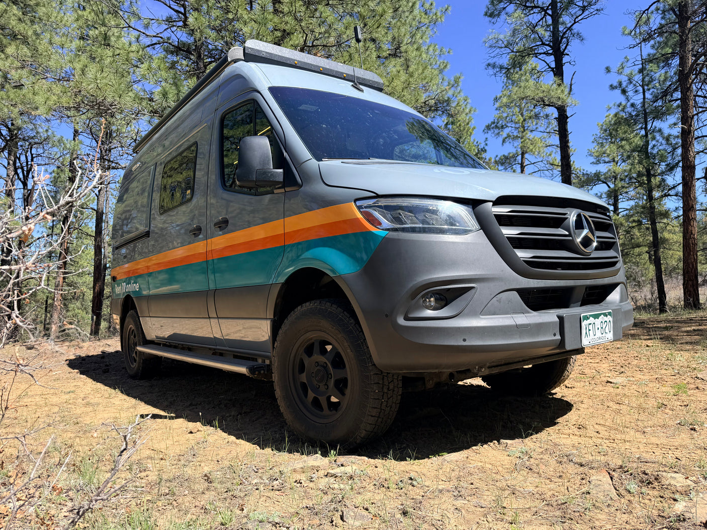 RV rental: Grey Mercedes Sprinter camper van with orange and teal stripes, parked in a pine forest.