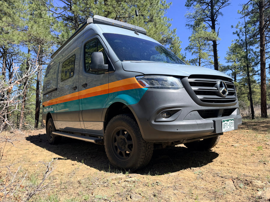 RV rental: Grey Mercedes Sprinter camper van with orange and teal stripes, parked in a pine forest.