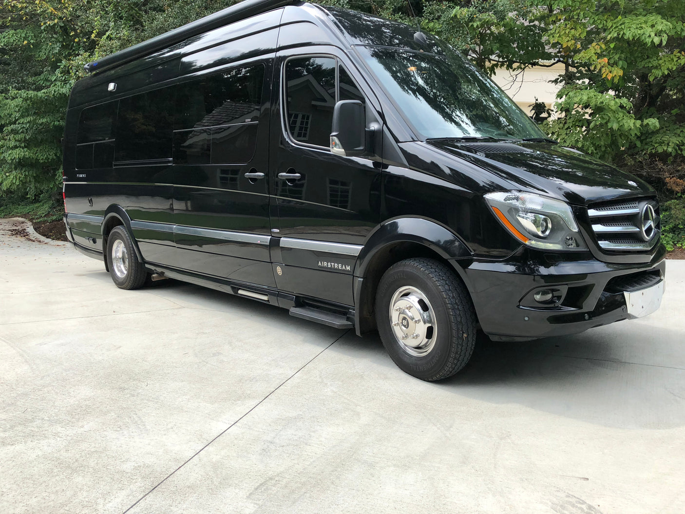 Black Airstream Mercedes Sprinter rv rental van, perfect for travel. Parked on a concrete driveway with trees in the background.