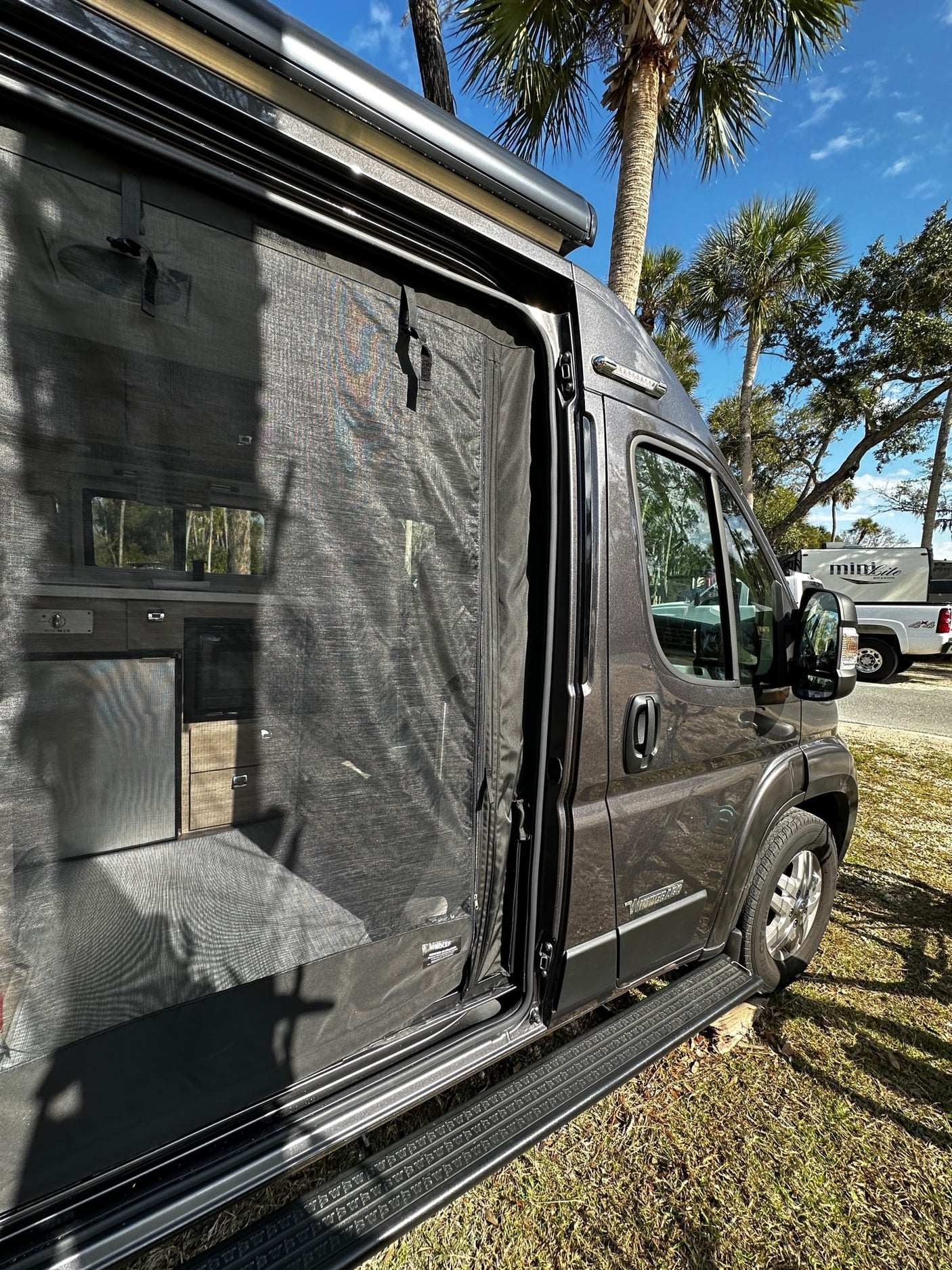 Adventure awaits with this rv rental! View of a dark RV's open screened door, revealing kitchen amenities and sunny outdoor surroundings.