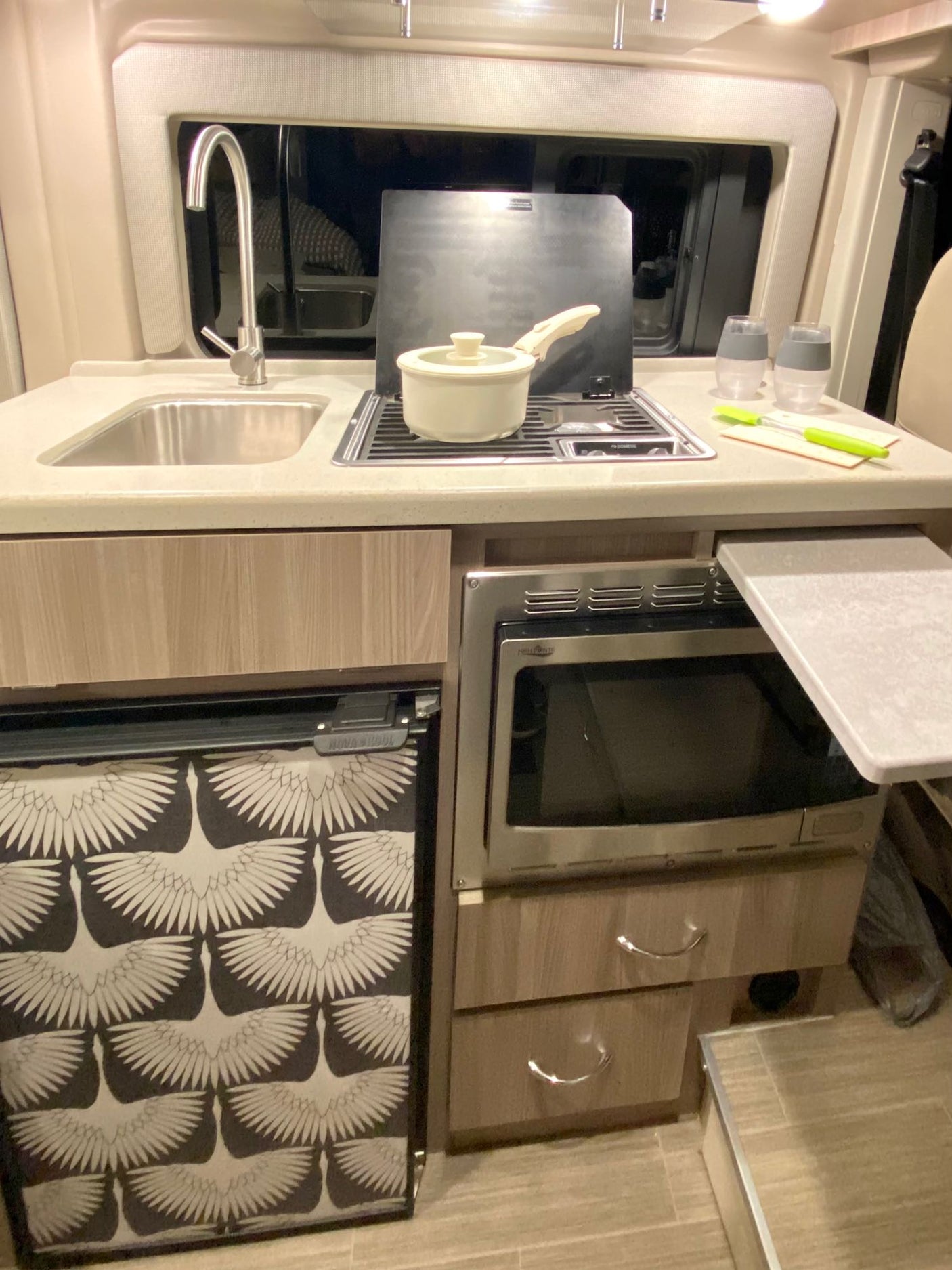 An RV rental kitchen shows a sink, stove, microwave, and uniquely patterned fridge, designed for efficiency.