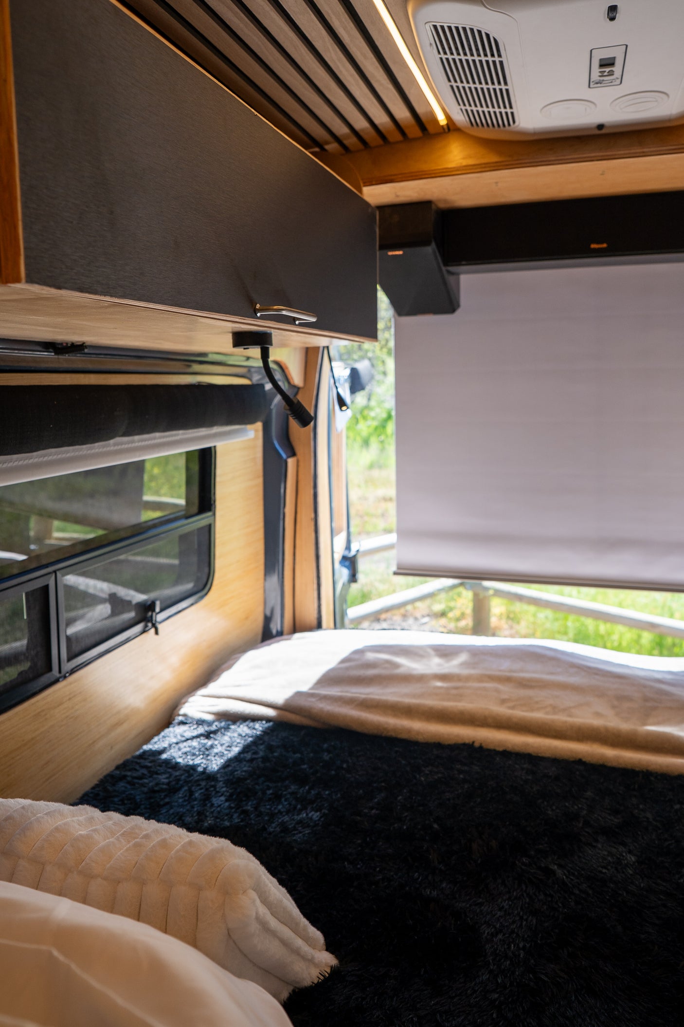 Inside an RV rental: a cozy bed with dark and white blankets, a window view, overhead cabinet, and AC unit.