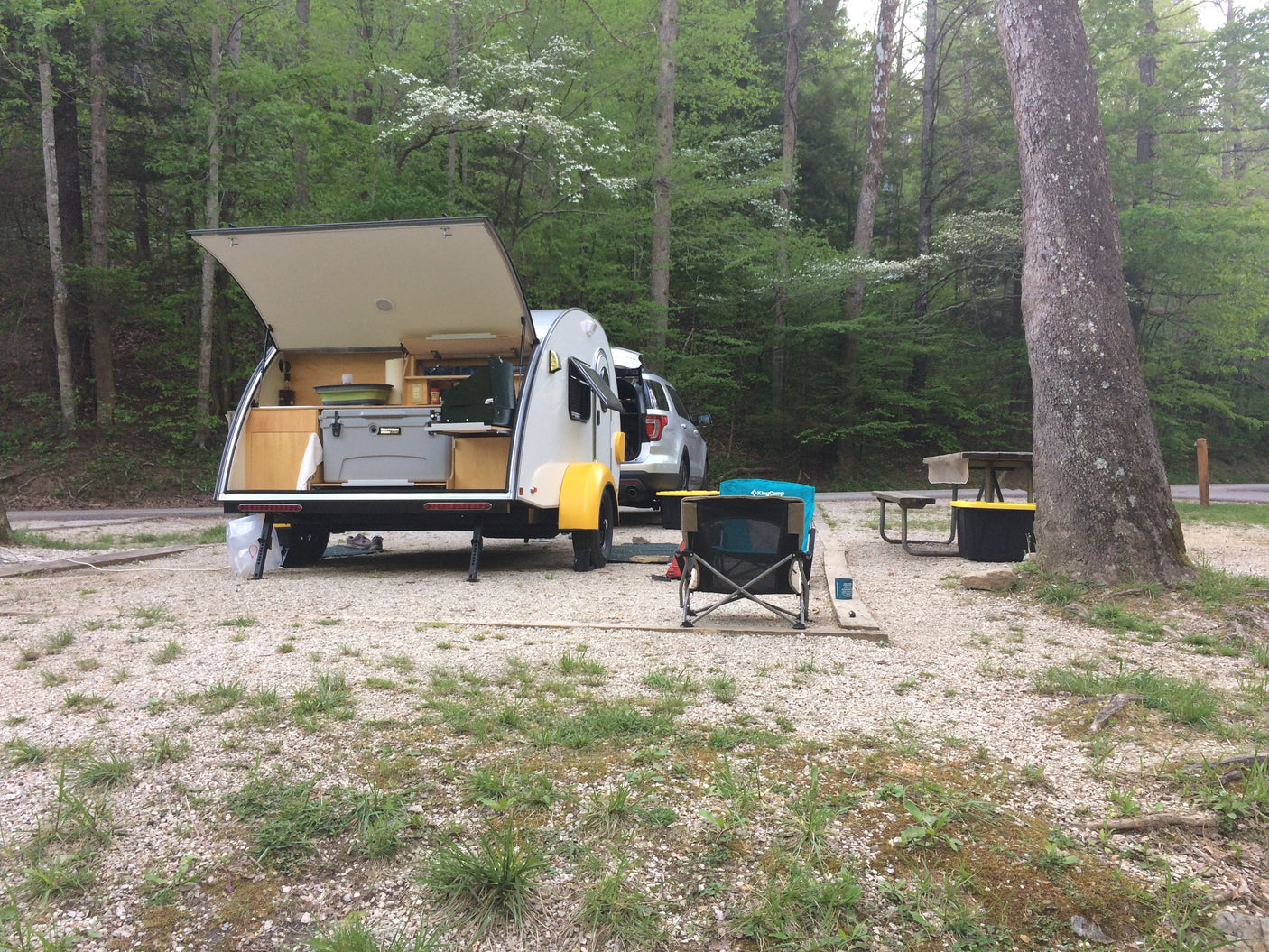 Teardrop RV rental ready for adventure at a gravel forest campsite, featuring an open kitchenette.