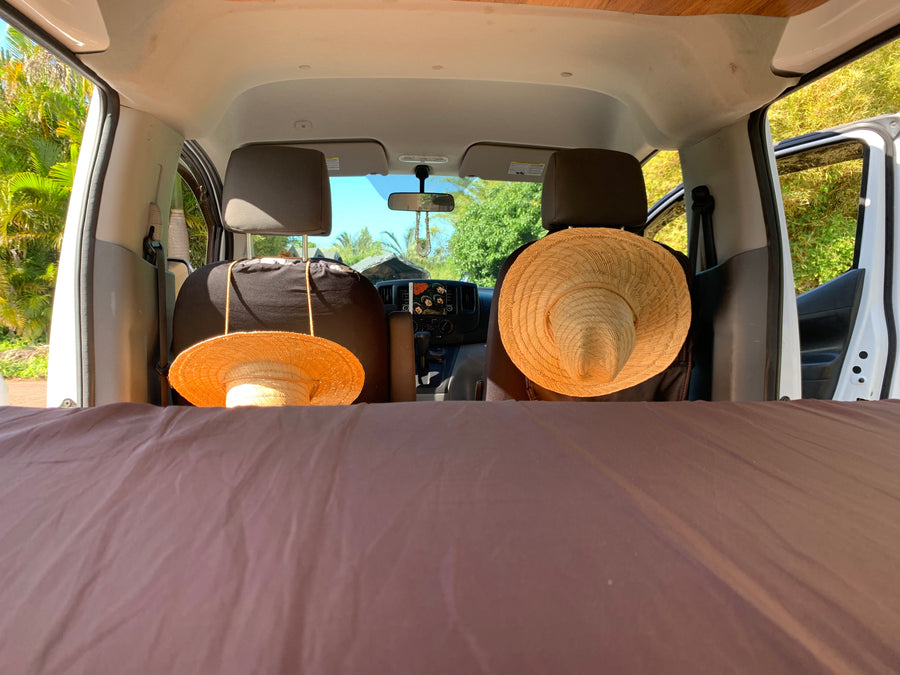 Your next RV rental adventure awaits! Cozy bed, hats on seats, and a tropical view from inside a camper van.