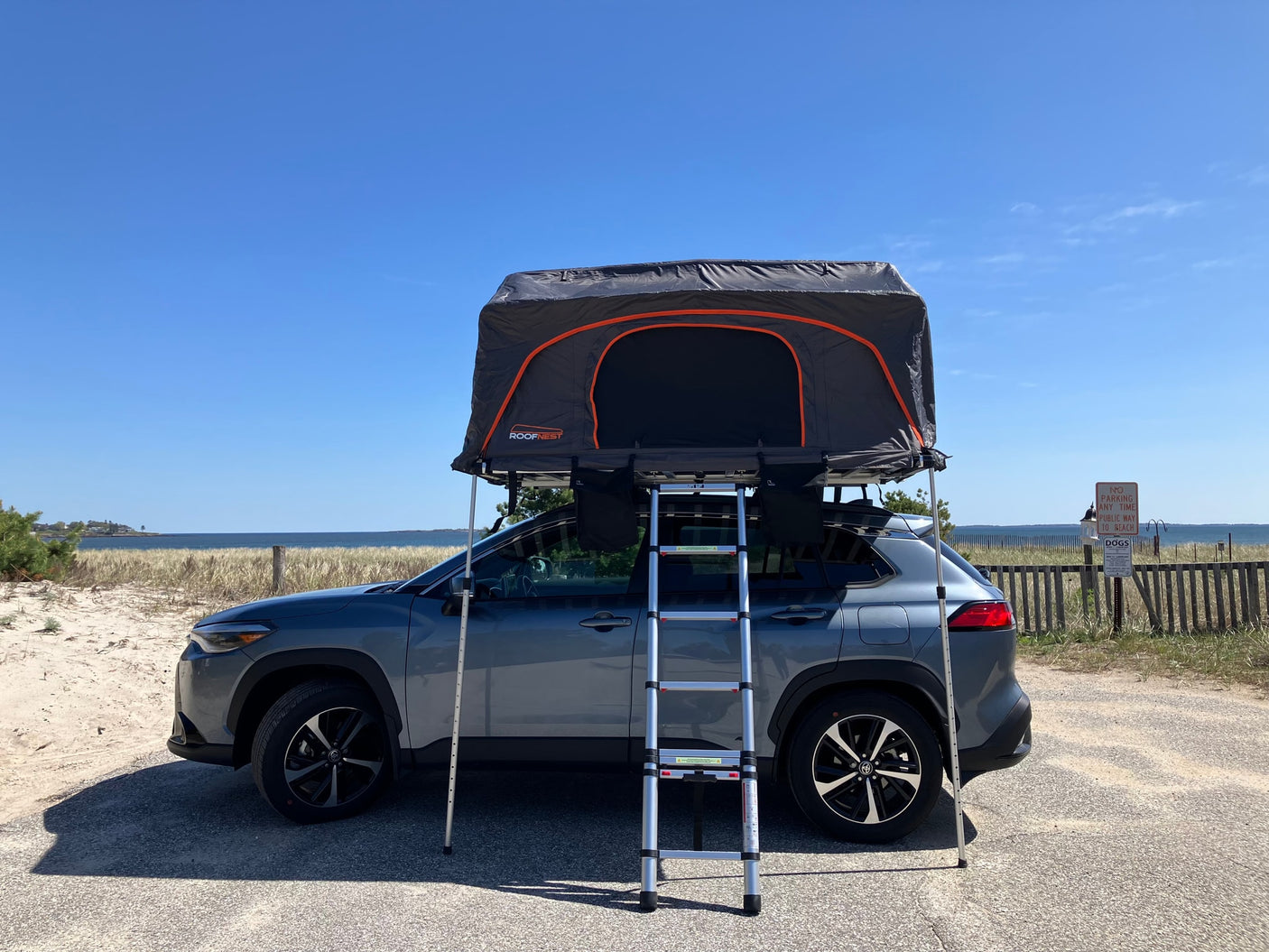 A gray SUV with a deployed rooftop tent stands by a sunny beach, ideal for your next RV rental camping adventure.
