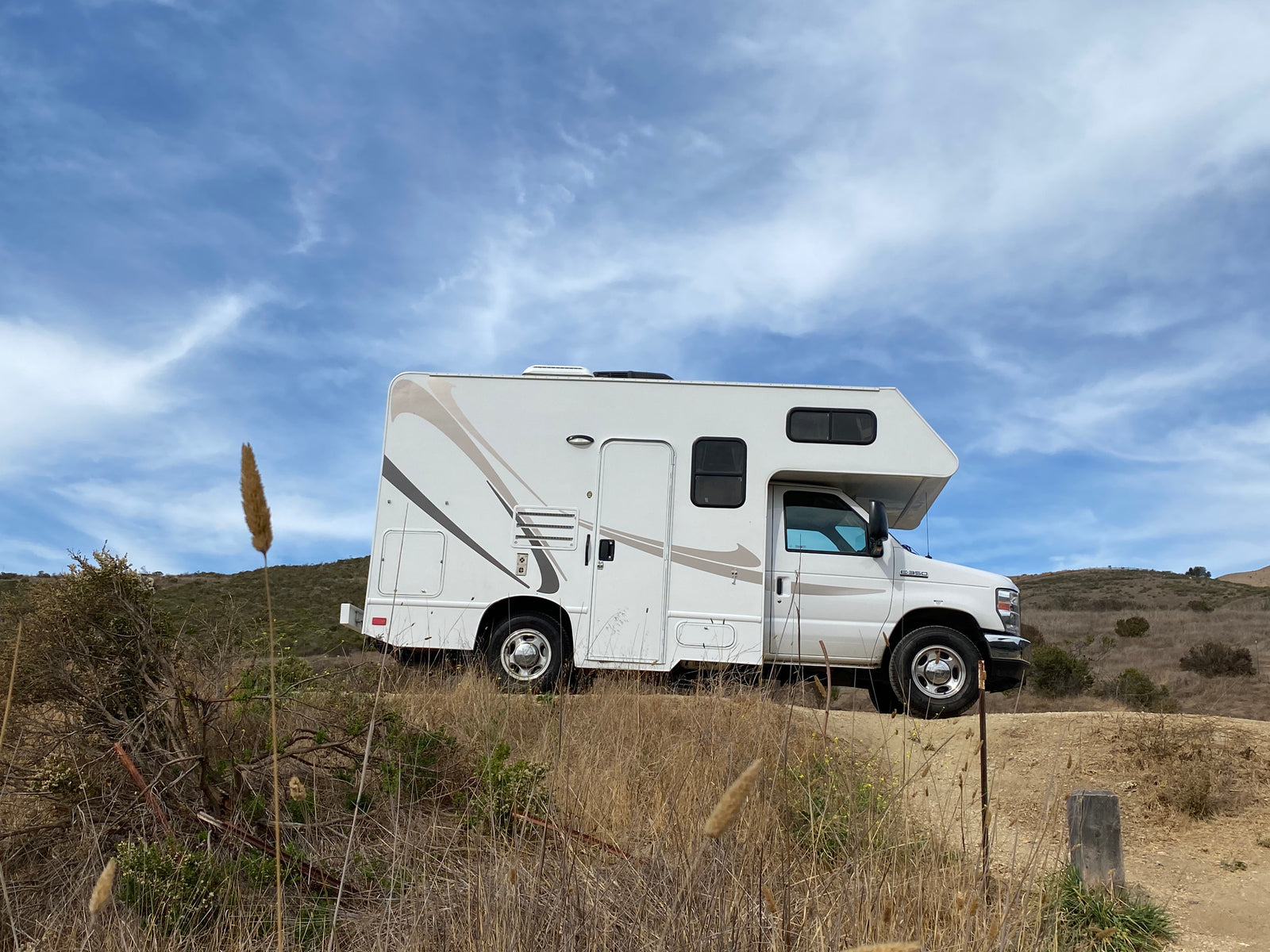 White RV rental parked on a dirt path in dry, grassy hills under a blue sky, perfect for outdoor adventures.