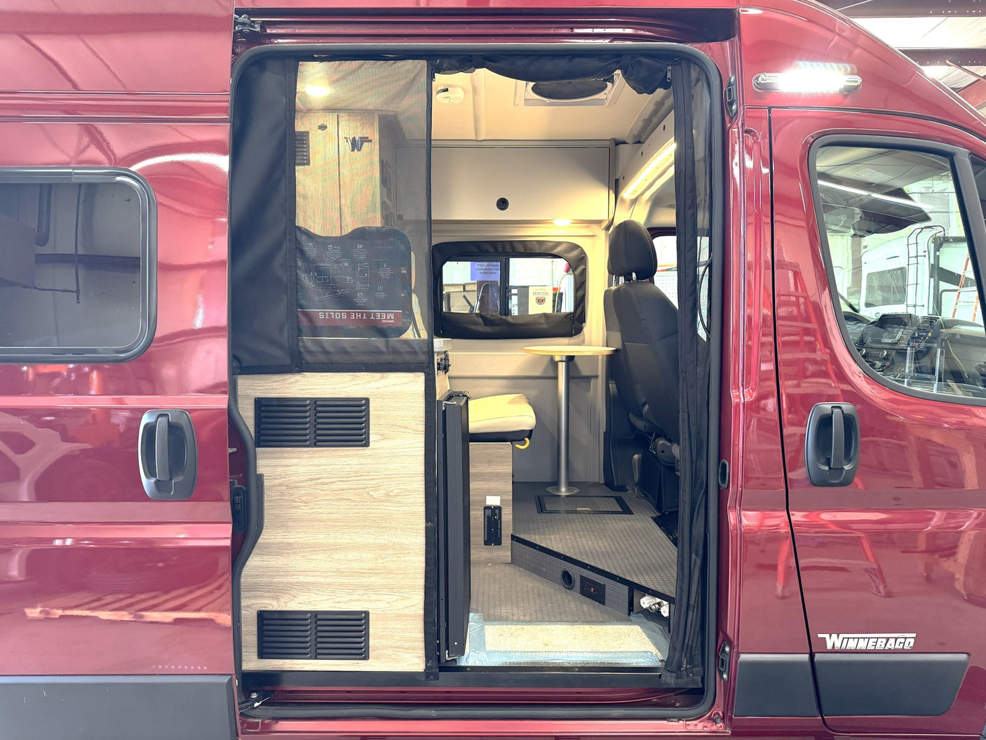 This red Winnebago camper van, perfect for rv rental, has an open side door revealing its compact interior with seating and table.