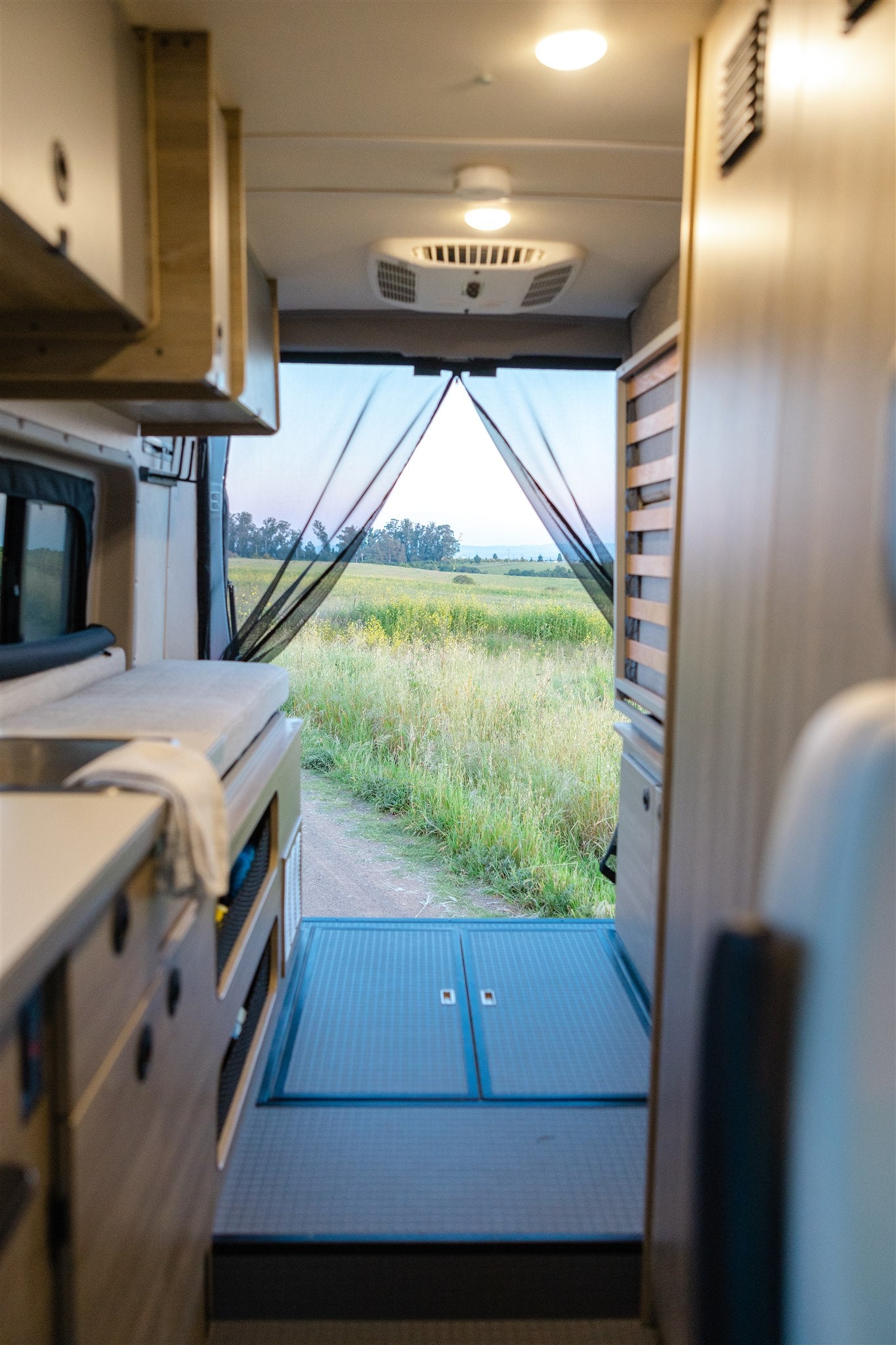 Your RV rental adventure awaits! Inside a camper van, overlooking a scenic grassy field with a path, protected by a mesh screen.