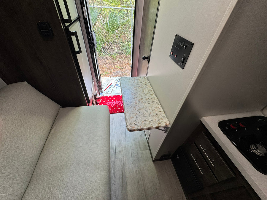 Inside an RV rental: light gray seating, wood-look floor, and a compact kitchen with a fold-down table near the open door.