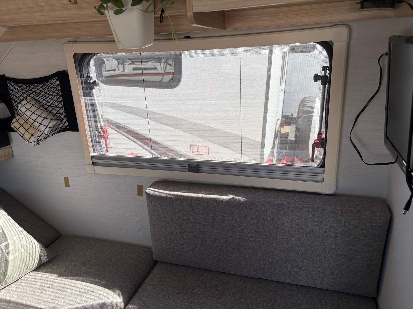 Cozy RV rental interior: bright window with screen, soft gray seating, storage net, and TV. Enjoy comfort on your road trip.