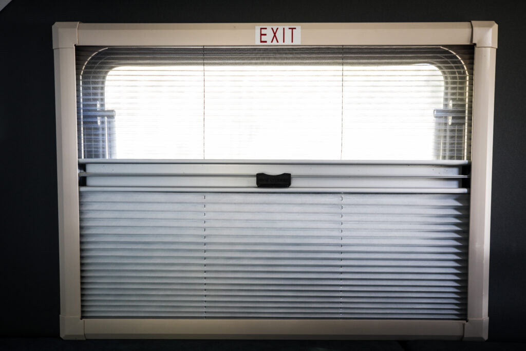 Inside an RV rental: a window with pleated blinds, partially open for light. An "EXIT" sign is above.