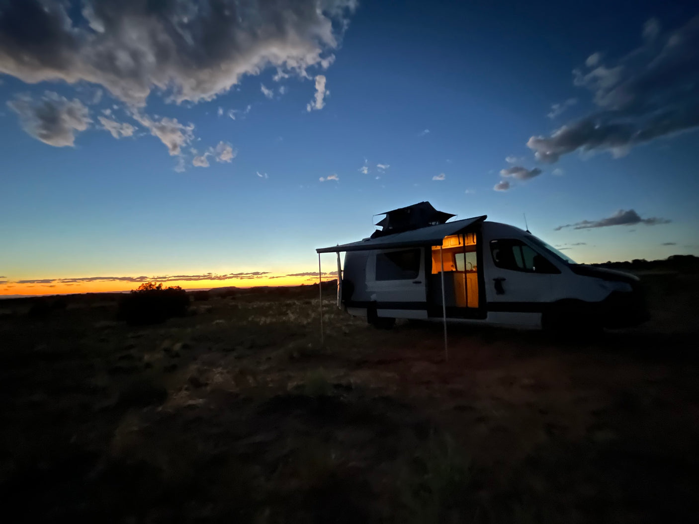 An RV rental parked at sunset in a vast desert. Interior lights glow, showcasing cozy amenities for off-grid camping.