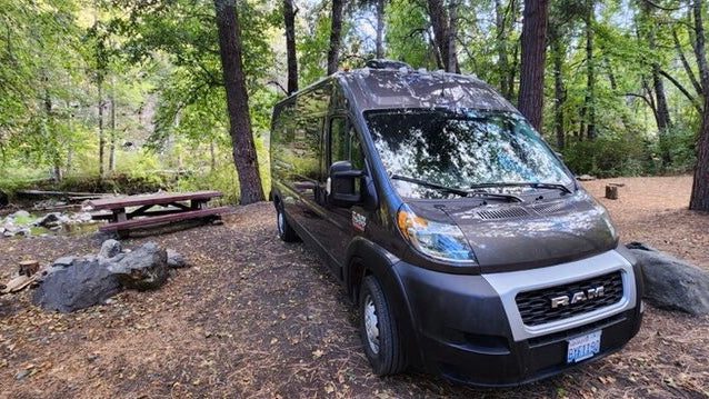 Dark gray camper van as an rv rental parked at a tranquil wooded campsite, with a picnic table & stream nearby.