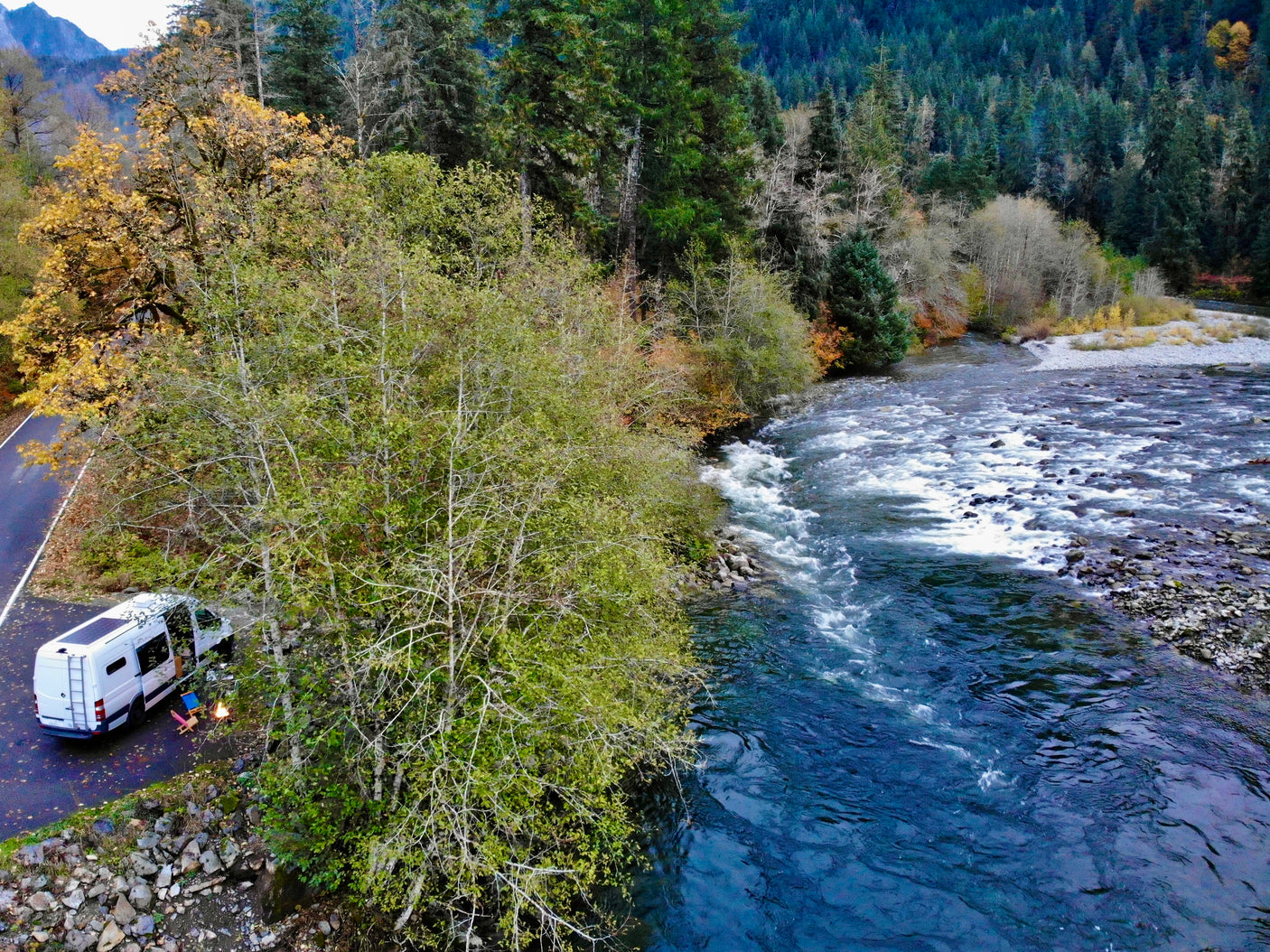 An RV rental is parked by a river, with a campfire and chairs, amidst an autumn forest. Perfect for scenic outdoor adventures.