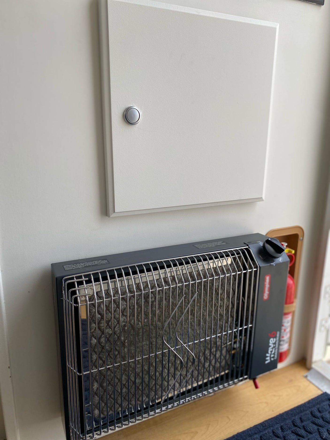 Compact Olympian Wave 6 propane heater and fire extinguisher inside an rv rental, with an access panel above.