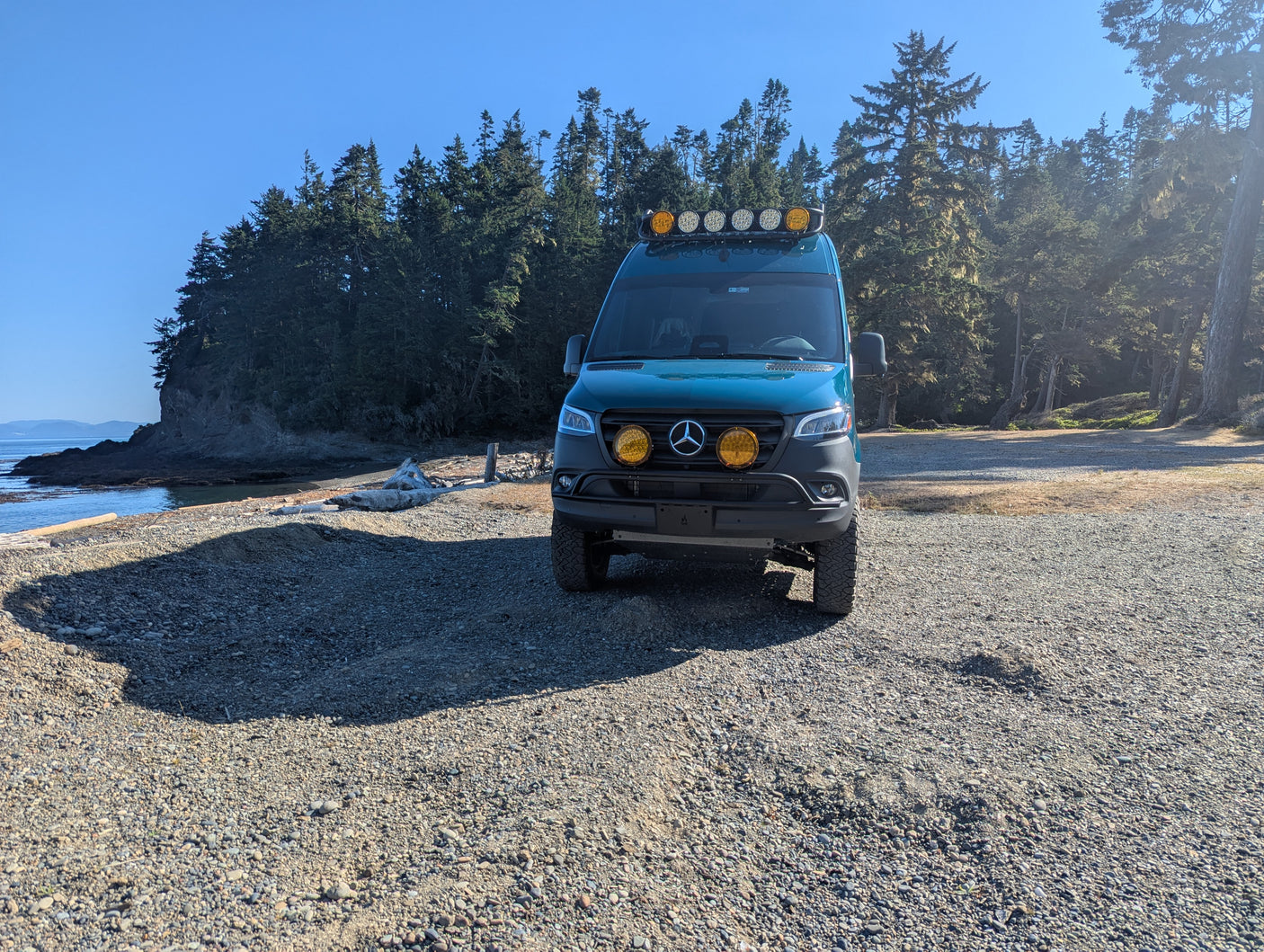Custom teal Sprinter van, perfect for an RV rental adventure, parked on a gravel beach near forested ocean cliffs.