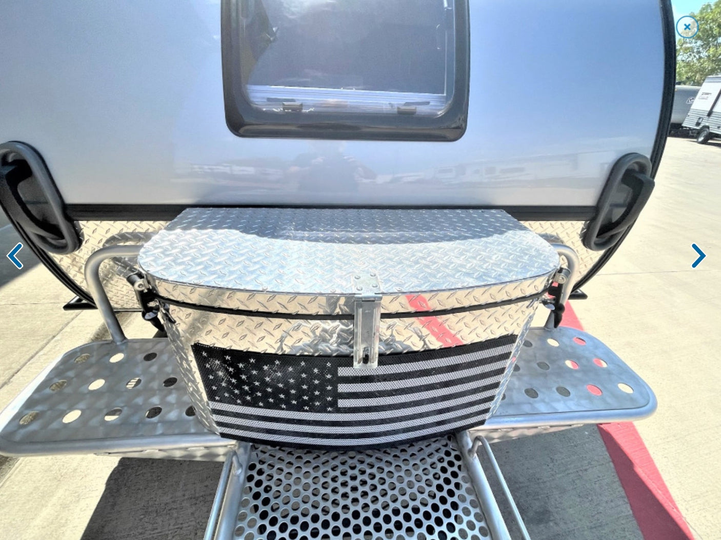 featuring front storage and roof rack.|Rear view of a tear-drop RV rental featuring a diamond plate storage box with an American flag design and perforated steps.|Explore off-grid adventures! This silver teardrop camper with a solar panel and spare tire is perfect for RV rental.|RV rental with a compact outdoor kitchen: sink