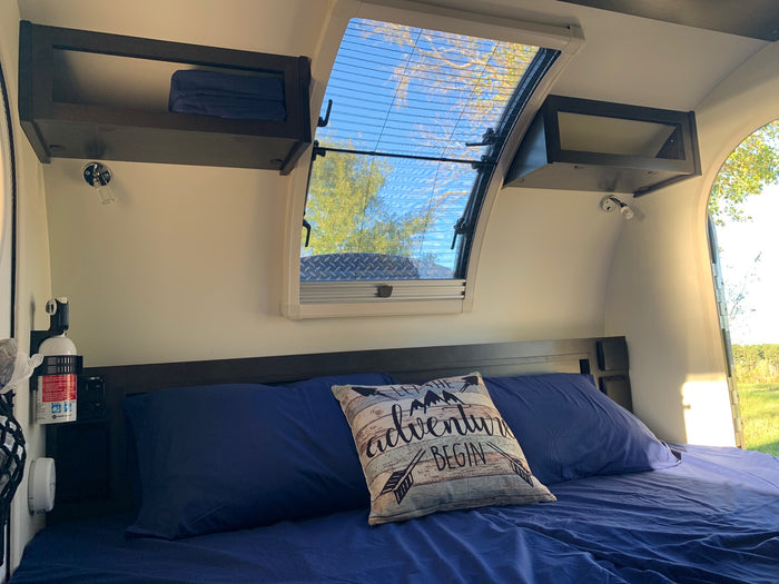 Cozy RV rental interior shows a blue bed, adventure pillow, overhead storage, & a bright skylight with a screen & outdoor view.