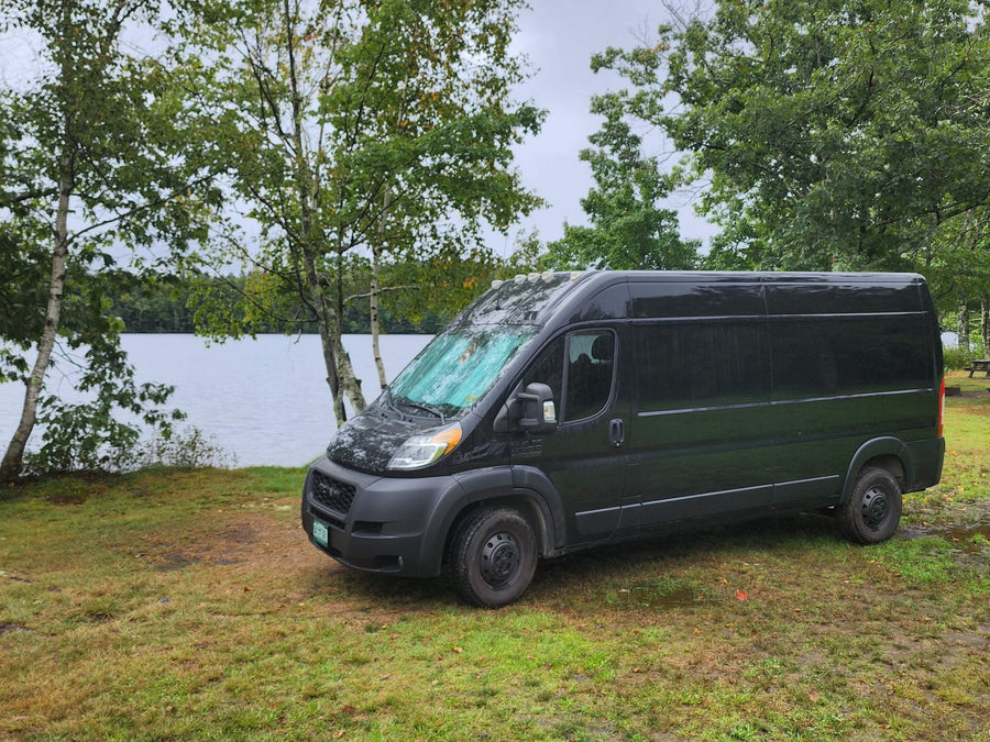 Serene lakeside view with a black RV rental van parked on damp grass under overcast skies. Explore nature's beauty.|Black Ram ProMaster van with bull bar & Vermont plate