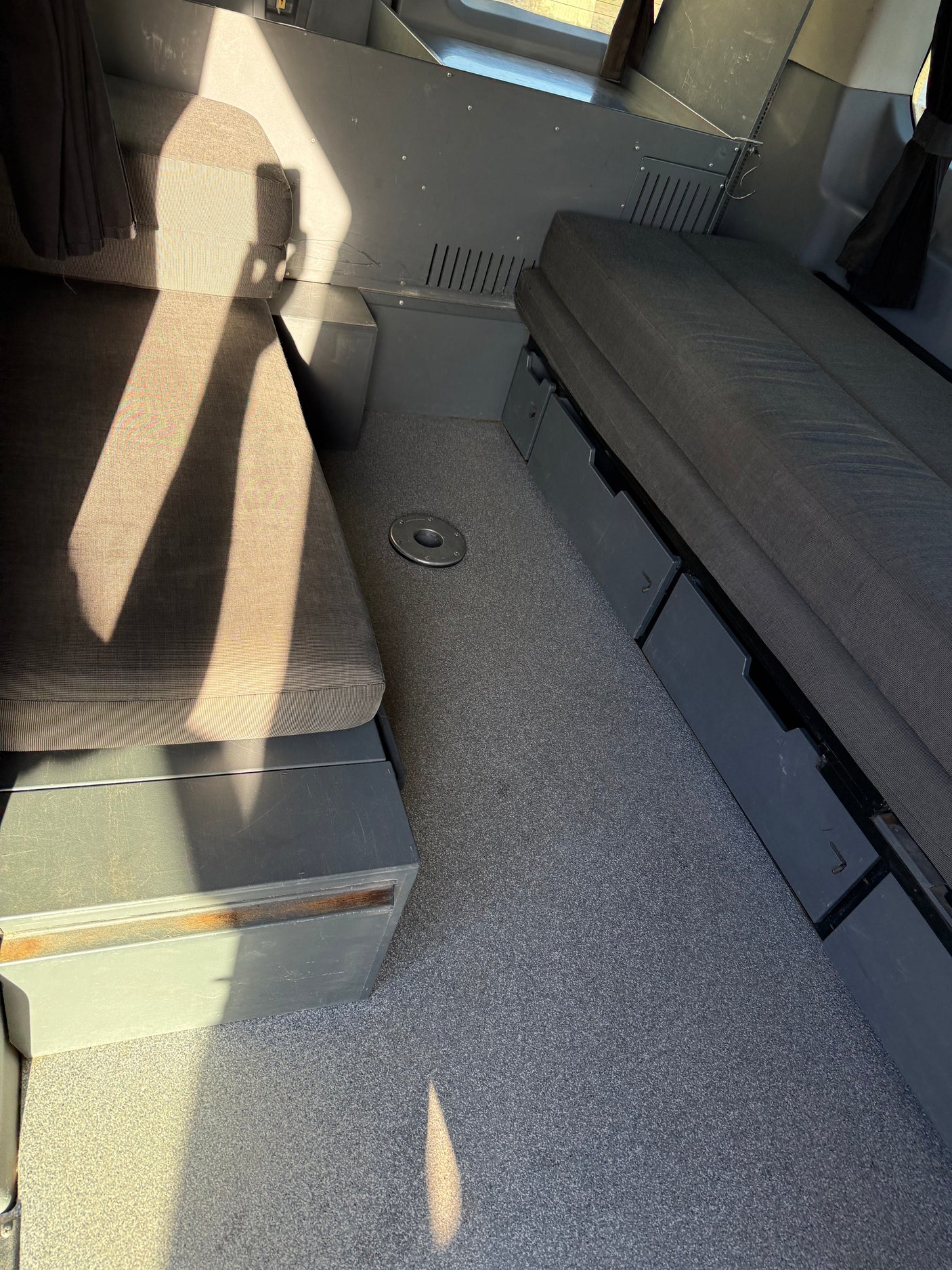 RV rental: Van interior showing cushioned benches, under-seat storage, table mount on grey carpet, sunlit.