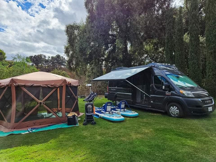 RV rental set up with brown screen tent & blue paddleboards on green grass, under cloudy skies.