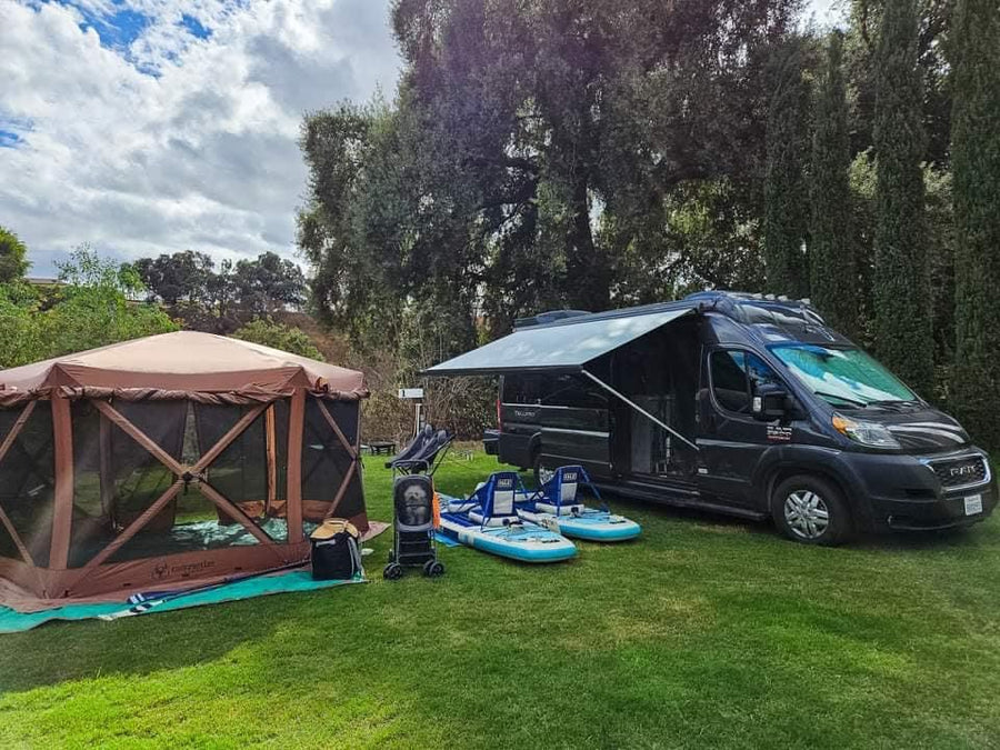RV rental set up with brown screen tent & blue paddleboards on green grass, under cloudy skies.