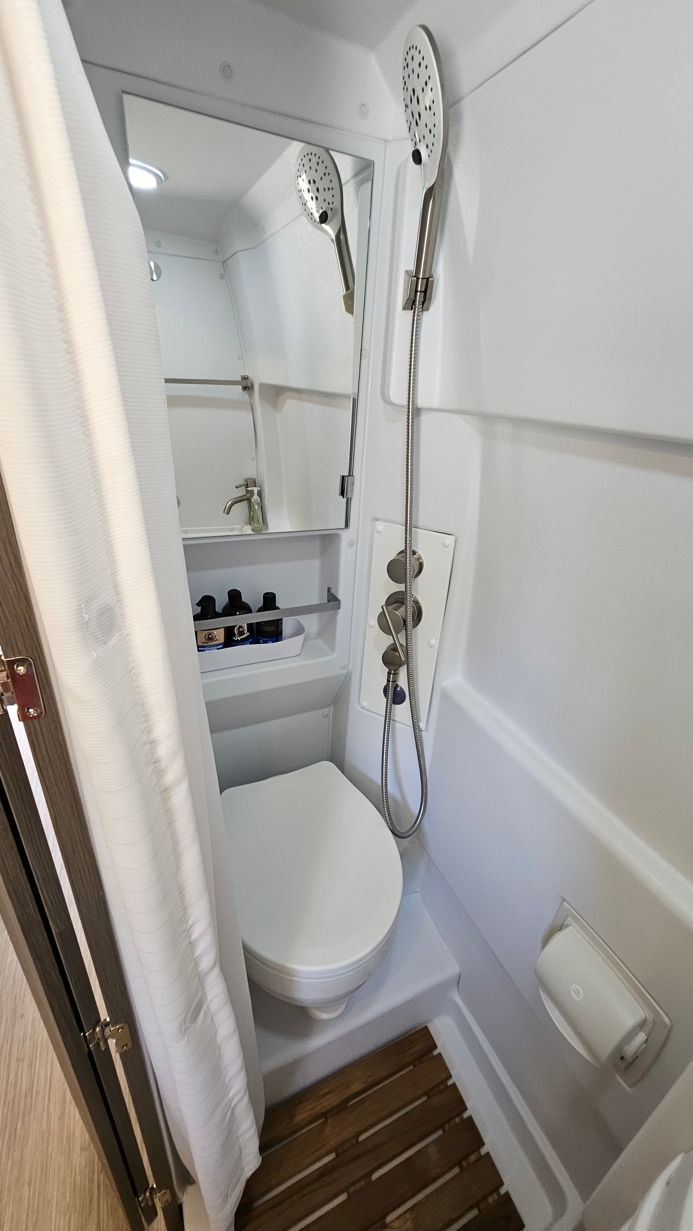 Compact RV rental wet bath with toilet, handheld shower, and sink (reflected in mirror). Features shower curtain & controls.
