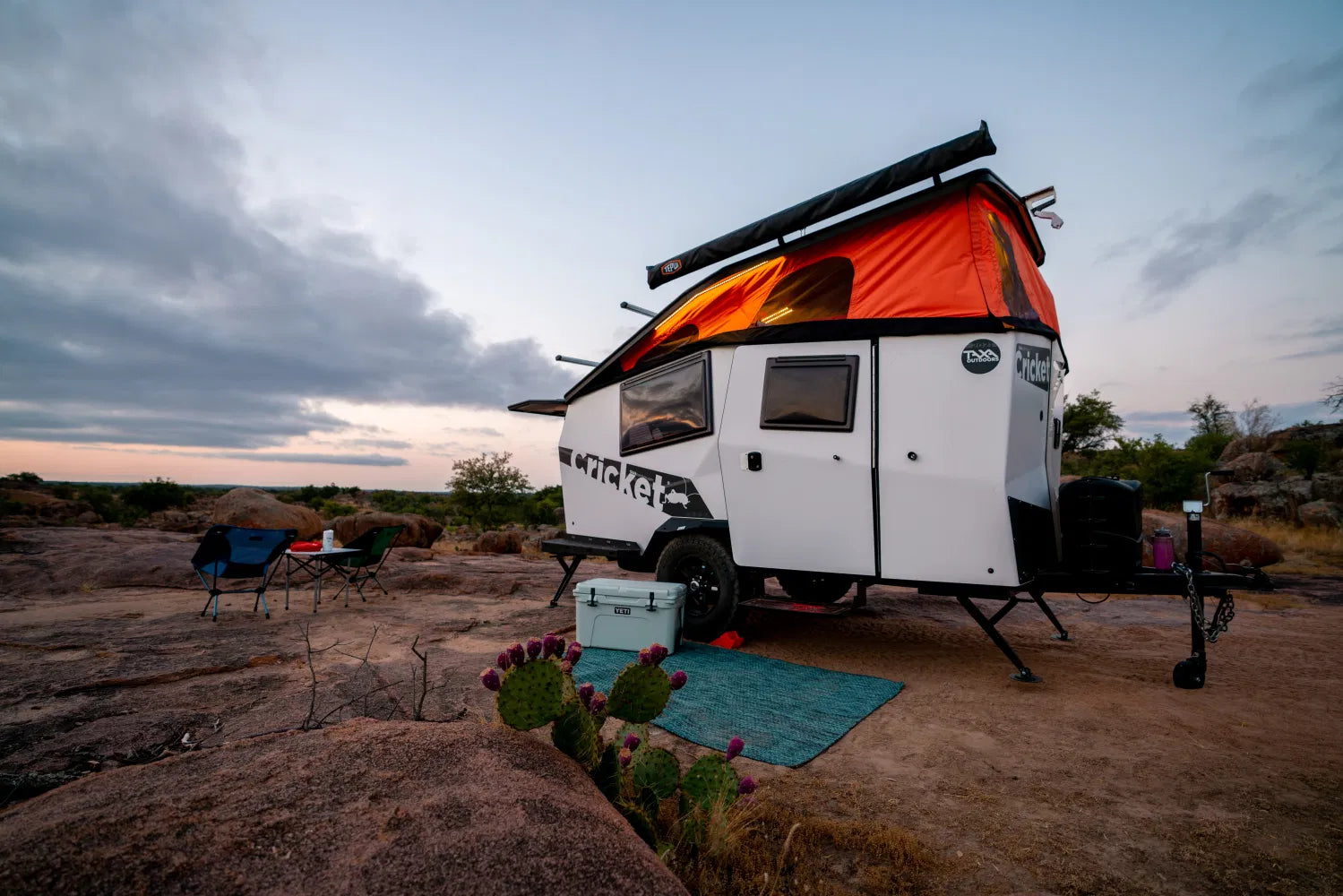 Experience outdoor adventure with an RV rental! A "Cricket" travel trailer set up for camping on a rocky landscape at dusk.|A rugged gray camper trailer
