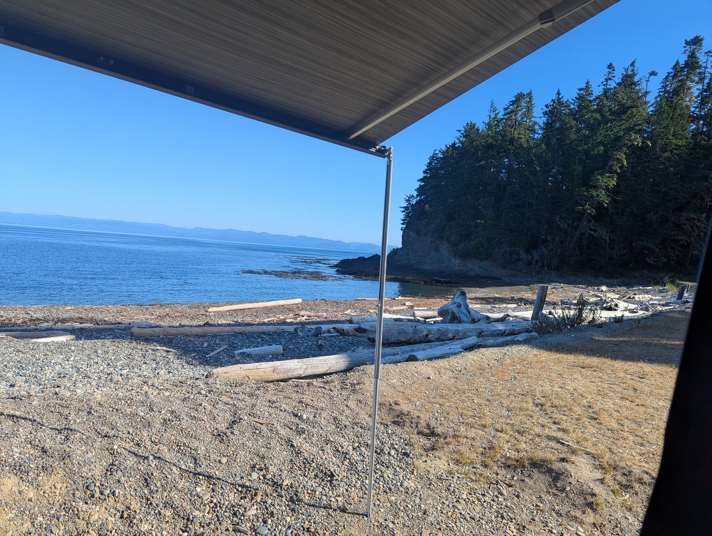 Enjoying a peaceful RV rental view of a sunlit beach, calm ocean, and forested coastline from under the awning.