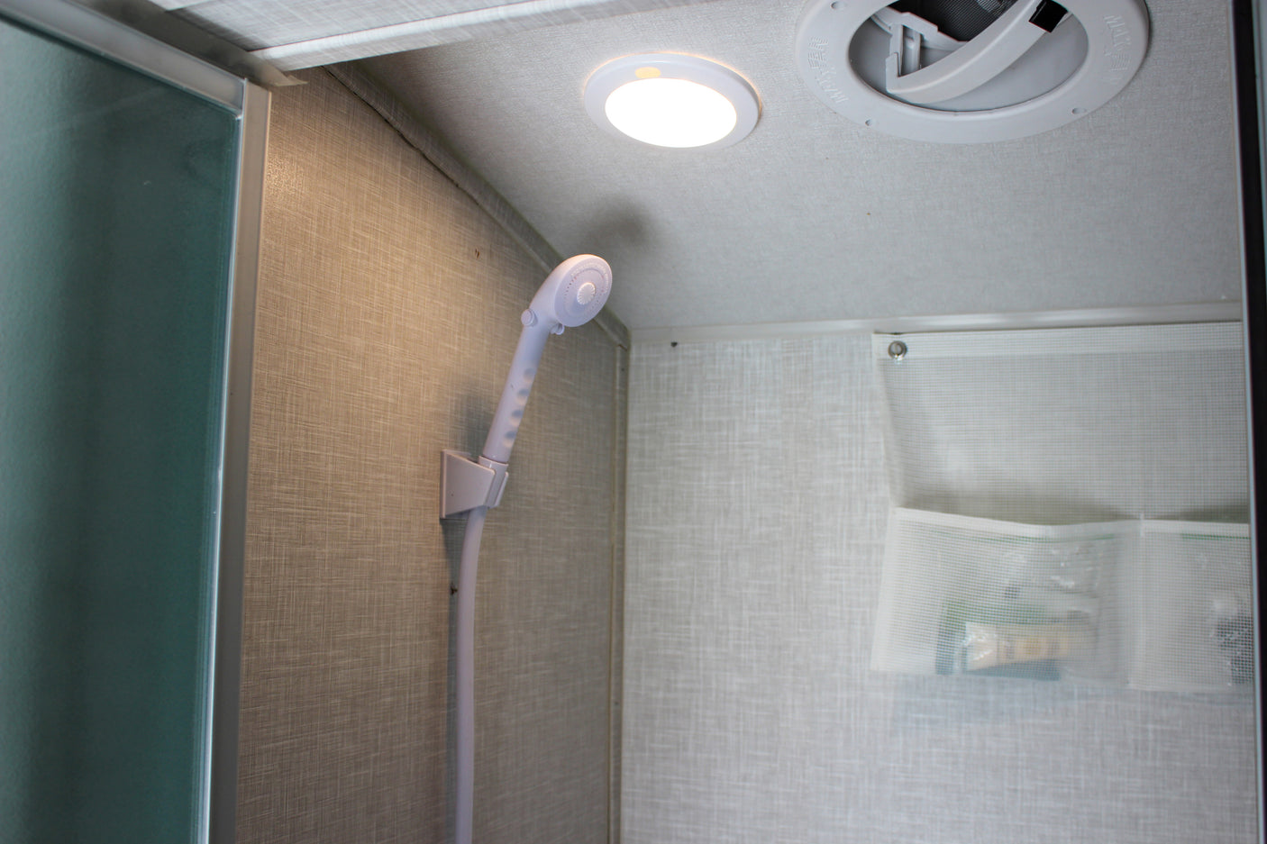 Compact RV rental shower interior: handheld shower head, ceiling light, ventilation fan, and wall-mounted mesh storage.