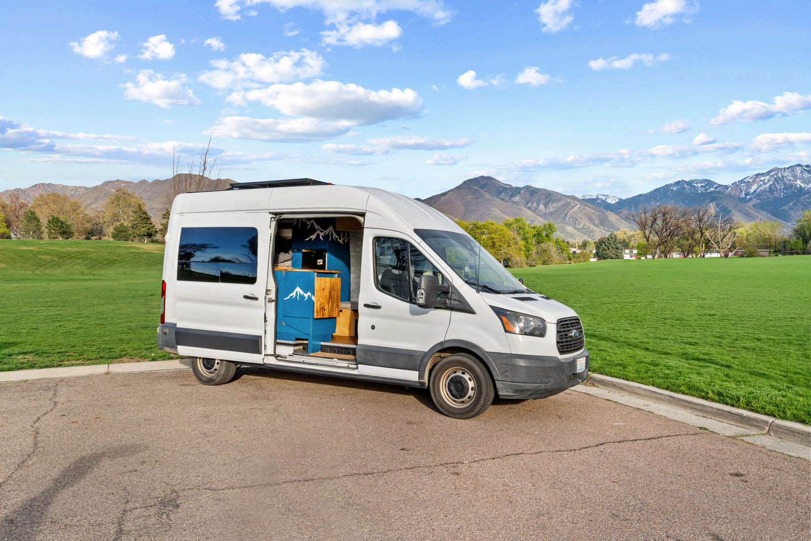 White Ford Transit camper van RV rental, with custom blue mountain-themed interior visible, parked by a green field with mountains in the background.