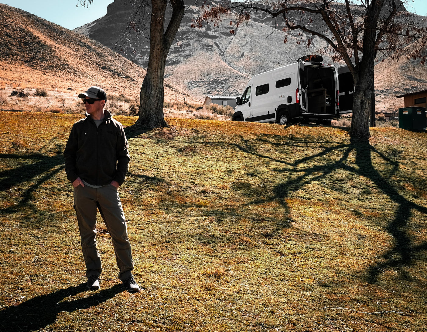 Scenic rv rental adventure. Man stands near his white camper van with open rear doors, parked in a grassy, mountainous landscape.