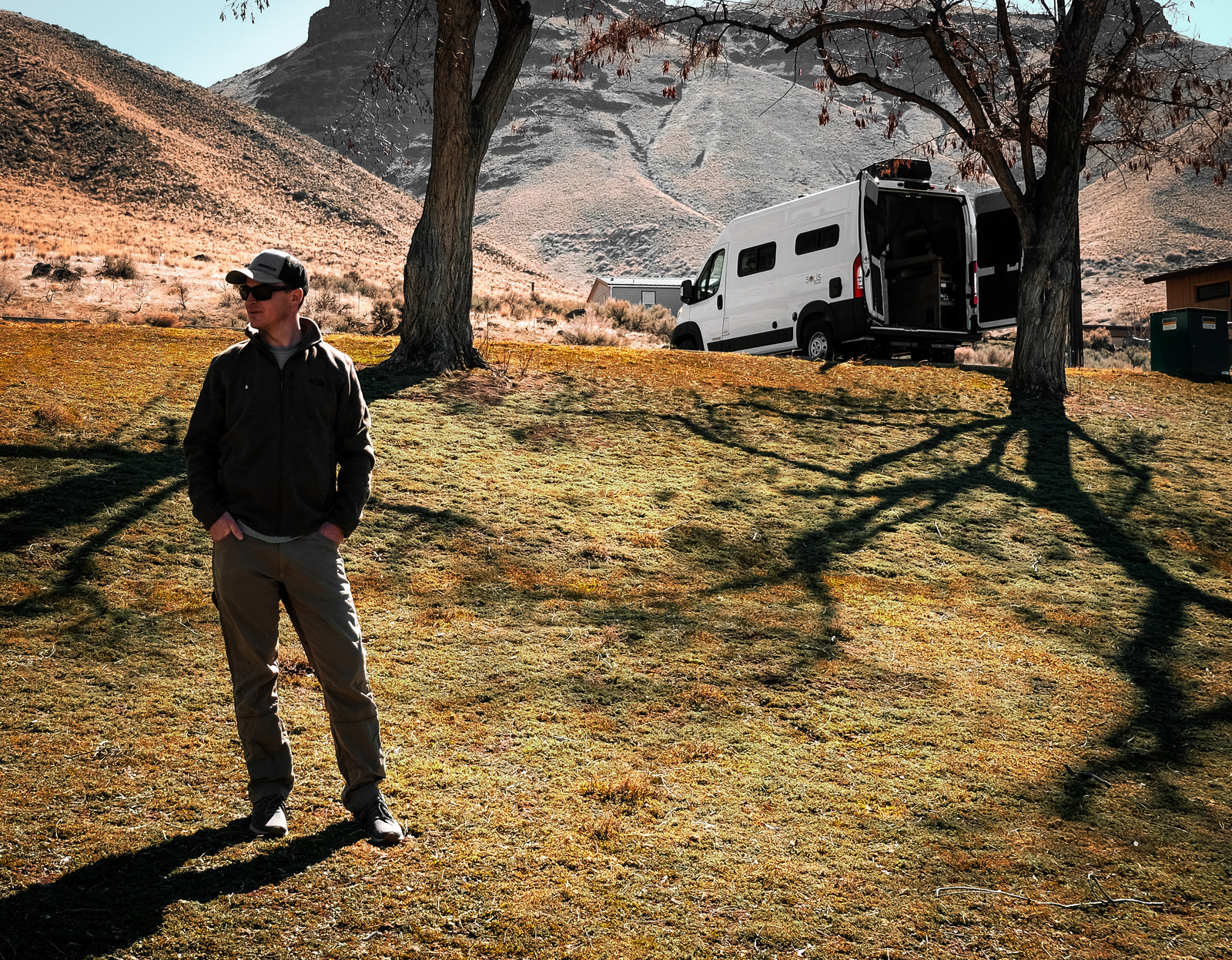 Scenic rv rental adventure. Man stands near his white camper van with open rear doors, parked in a grassy, mountainous landscape.