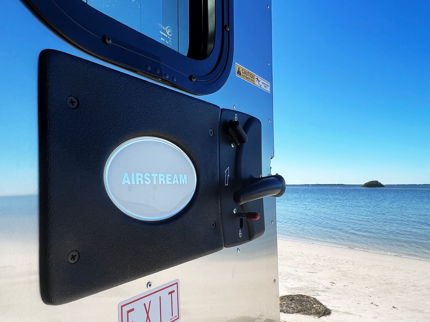 Airstream RV rental door with visible logo, handle, and caution sticker, facing a beach with clear blue water and sky.