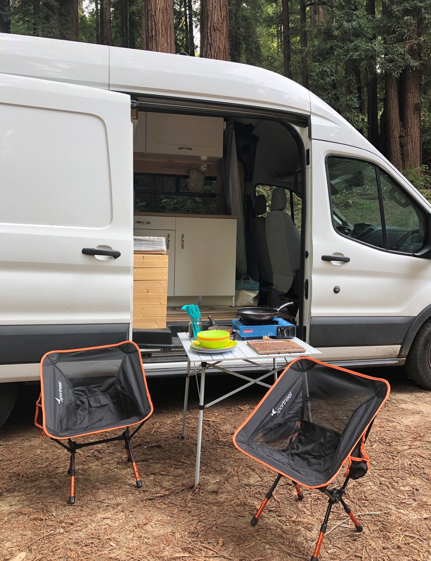 White camper van RV rental set up for outdoor cooking & dining. Portable stove, table, and chairs in a serene forest setting.