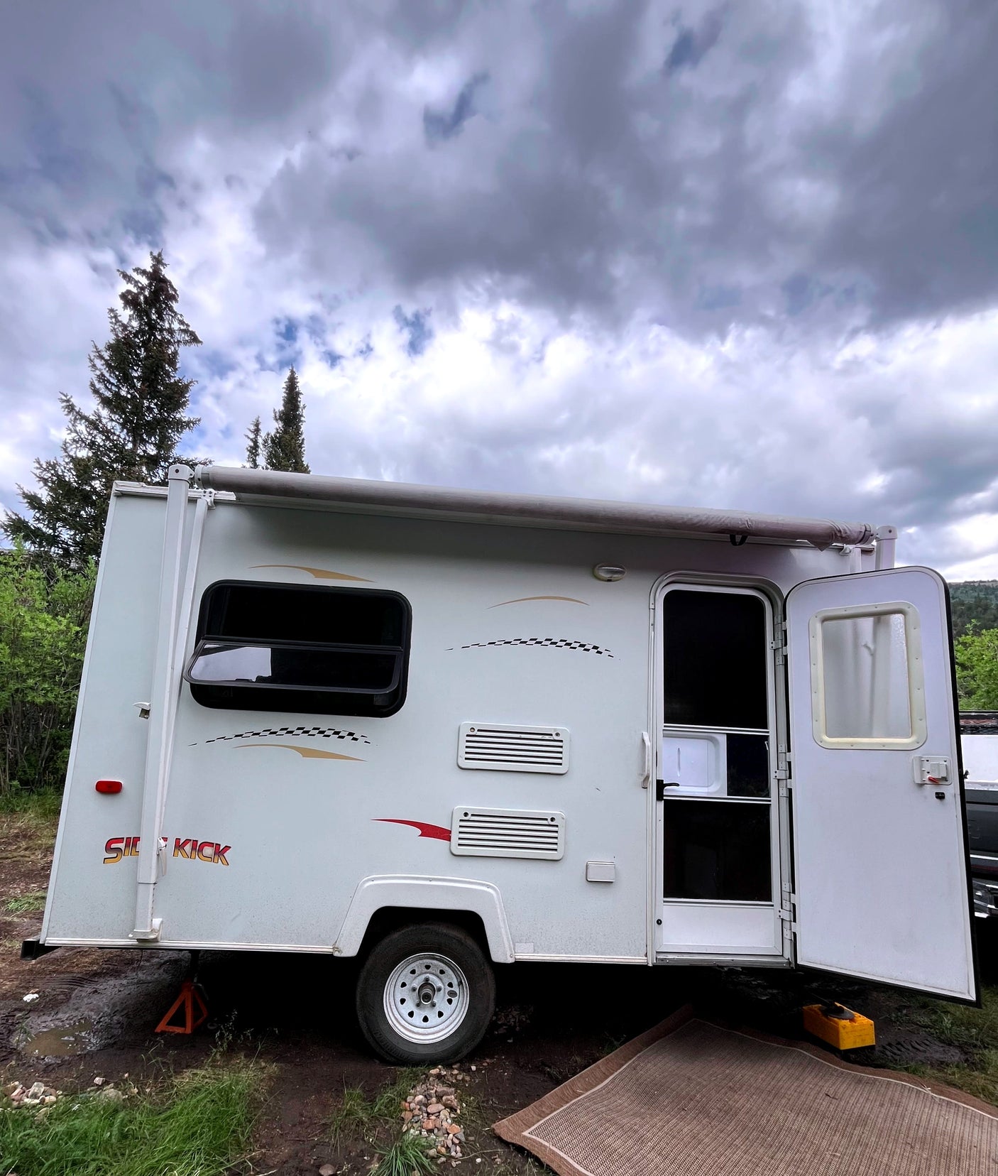 White travel trailer rv rental parked on muddy ground with an open door, surrounded by trees under a cloudy sky.