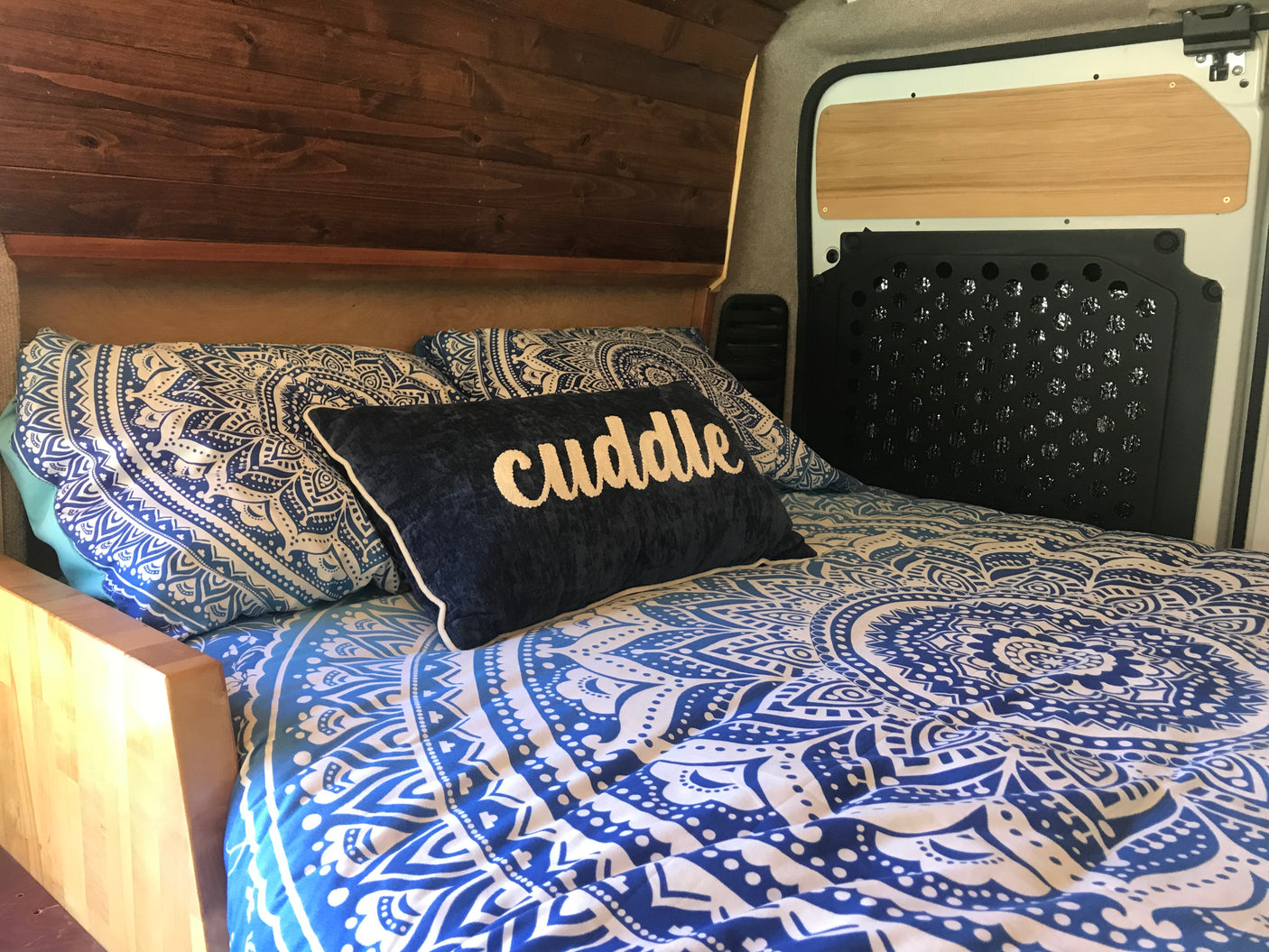 Cozy RV rental bed with blue mandala bedding and "cuddle" pillow. Features warm wooden interior and details.