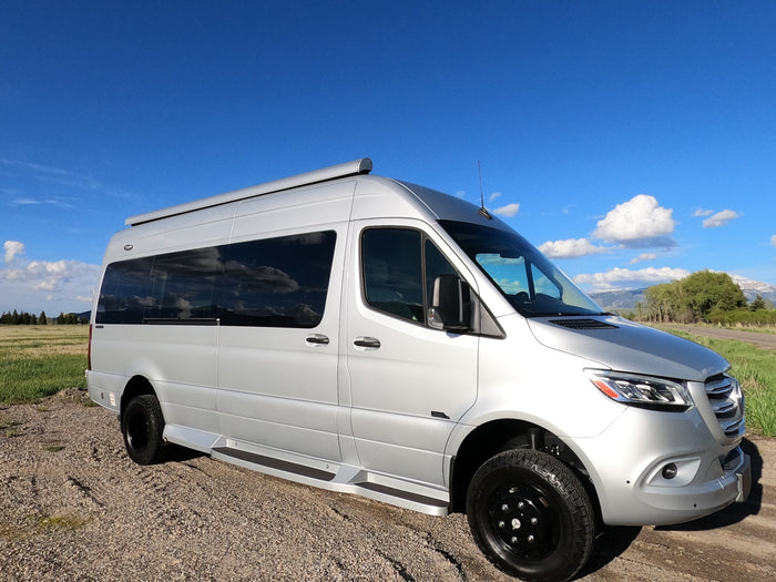 This sleek silver rv rental van is ready for adventure, parked on a gravel path amidst fields and distant mountains.