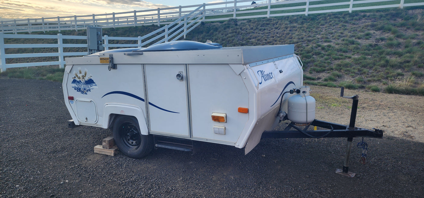 A white Aliner Sportliner pop-up RV camper parked on gravel with a fence and grassy hill behind. Perfect for your rv rental!