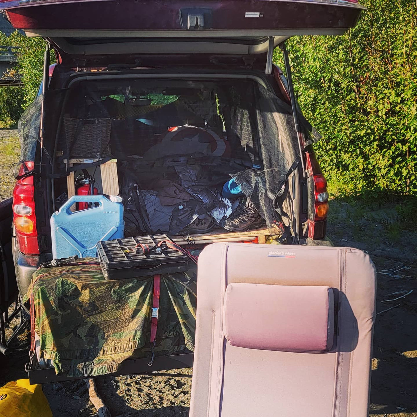 Car camping setup: open tailgate reveals packed gear inside a vehicle, with water jug & cot outside. Ideal for an RV rental alternative.