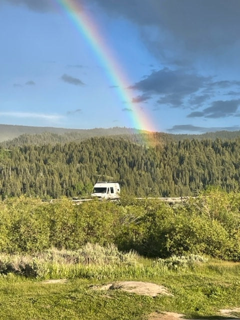 A vibrant rainbow arcs over a forest, with an RV nestled in the green foreground. Explore nature's beauty with an RV rental. — photo 10