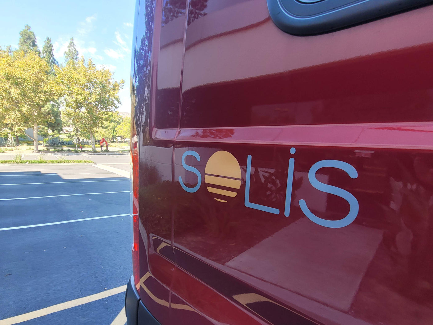 Red Solis RV rental van with its distinctive sun logo, parked outdoors on a sunny day near trees.