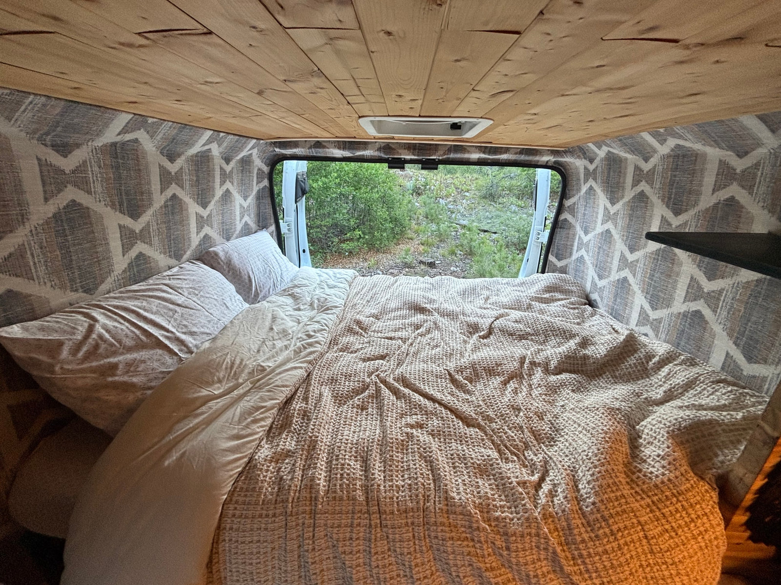 Cozy RV rental interior featuring a comfortable bed, wooden ceiling, patterned walls, and a beautiful nature view.