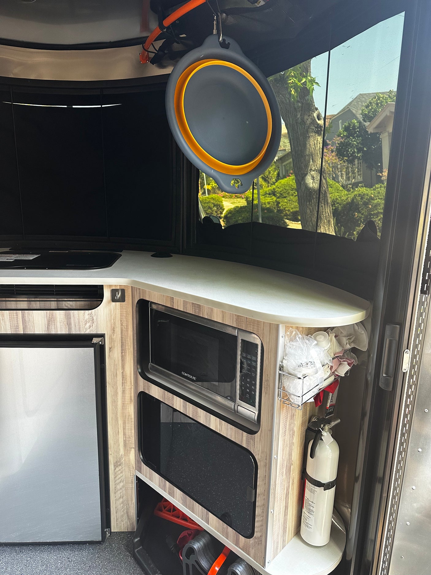 Inside RV rental: compact kitchen with microwave, fridge, cooktop, and hanging collapsible bowl. Fire extinguisher visible.