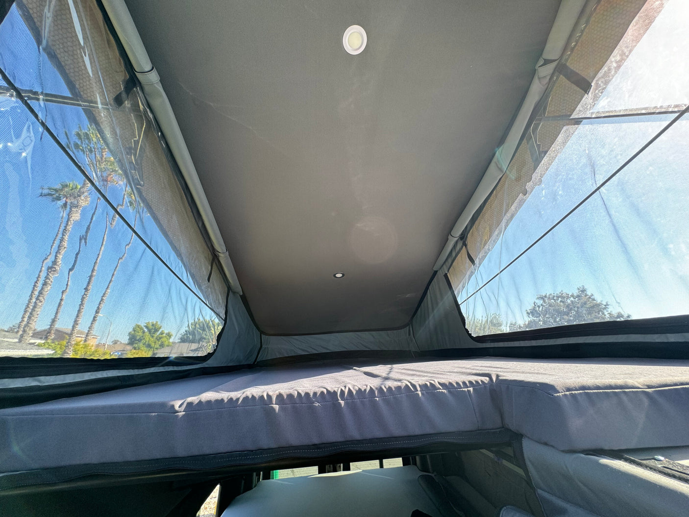 Spacious rv rental pop-top camper interior with a cozy bed, ceiling lights, and clear window views of palm trees and sky.