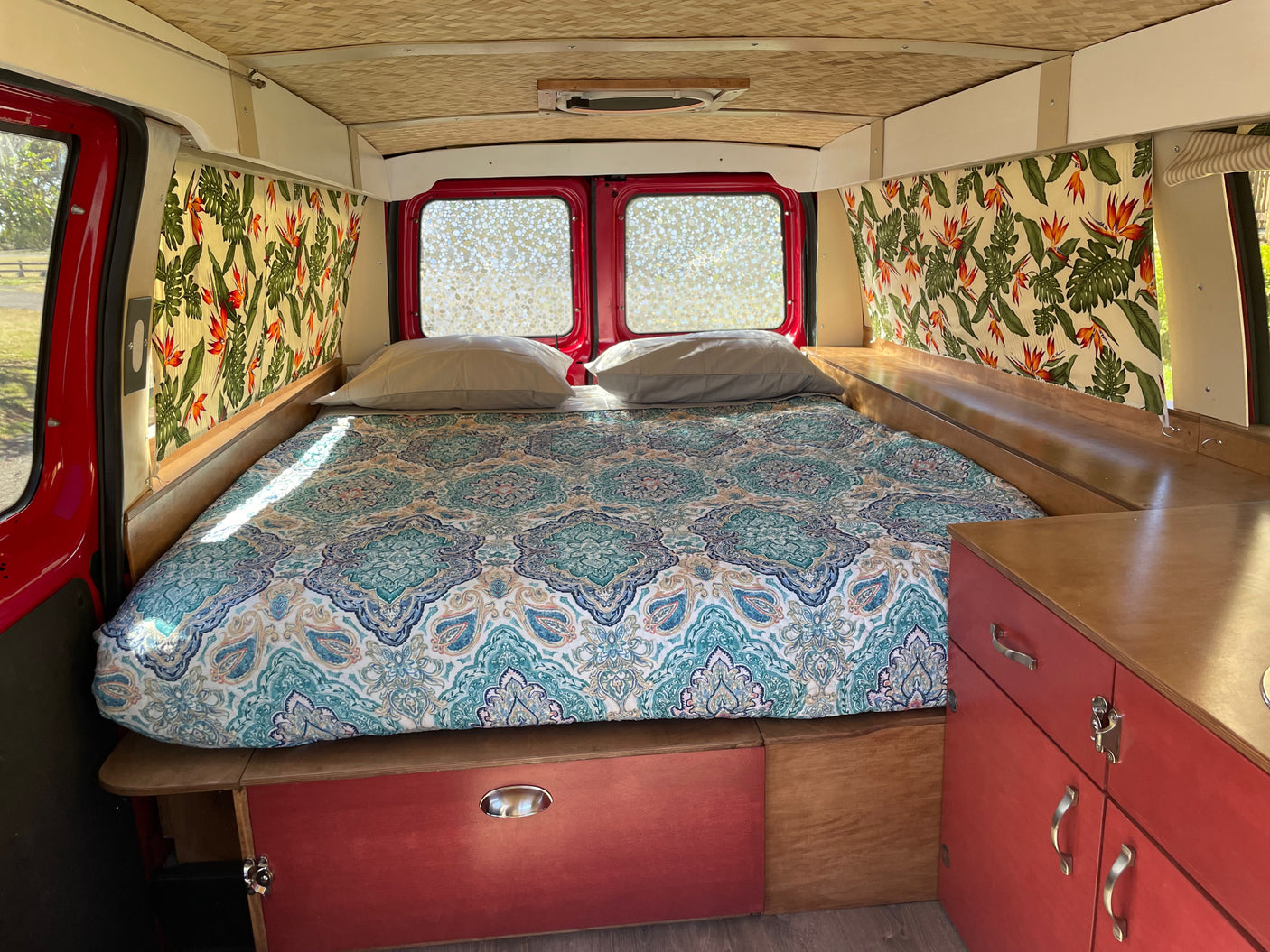 Inside this cozy RV rental: a comfortable bed with tropical print curtains and red cabinets, ideal for travel.