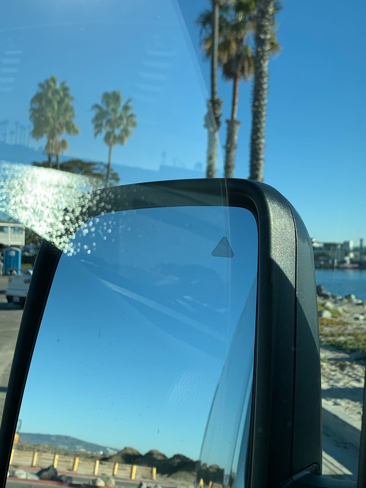 RV rental view: side mirror with blind spot monitor, palm trees & ocean under blue sky.