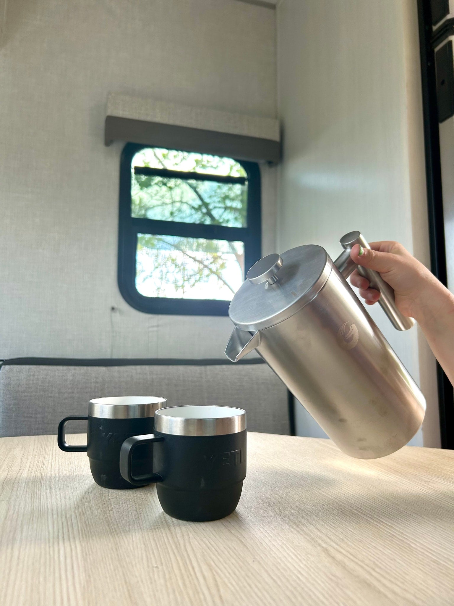 Coffee time in an RV rental! A hand pours from a stainless steel French press into two black YETI mugs on a light wood table.