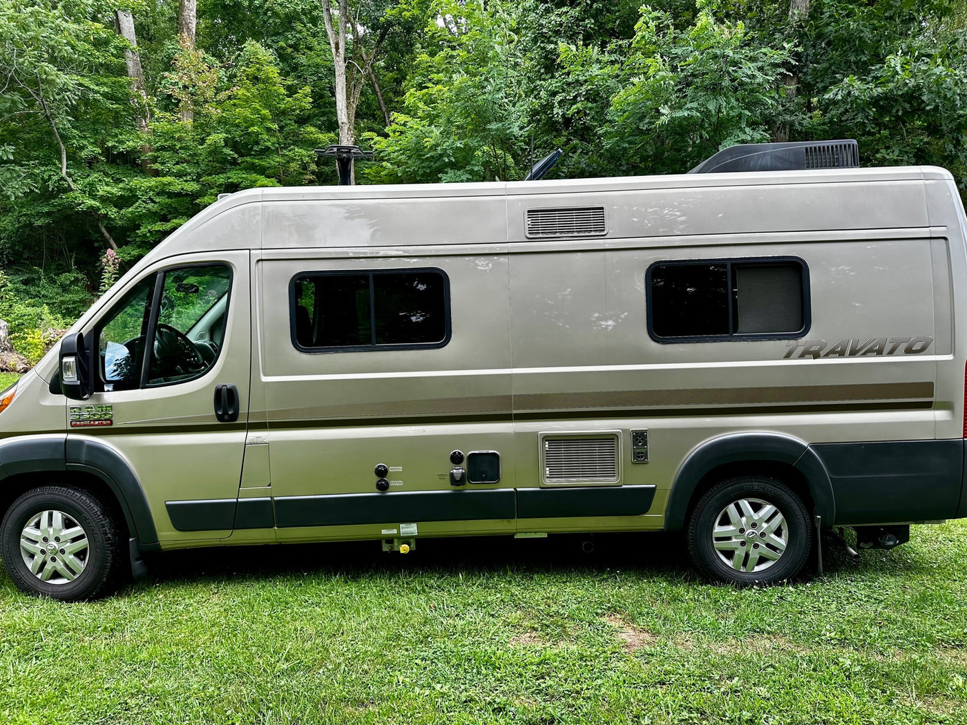 a comfortable rv rental option parked on green grass. Ideal for travel.|RV rental: Tan Ram ProMaster Travato camper van parked on green grass in a wooded area — photo 3